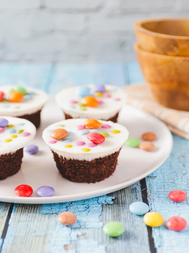 Ein weißer Teller mit vier Schokoladen-Cupcakes, garniert mit weißem Zuckerguss und bunten Bonbons. Verschiedene bunte Bonbons sind auf der blauen Holzoberfläche rund um den Teller verstreut. Im Hintergrund stehen drei gestapelte Holzschüsseln auf einem Tuch vom Food Fotograf Hannover.