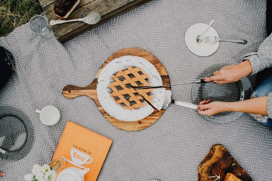 Food Fotograf Eine Person schneidet einen Kuchen mit einem Messer und serviert ihn mit einem Spatel auf einem Holzbrett. Das Brett steht auf einer grauen Picknickdecke, umgeben von Tellern, Besteck, einem Buch mit dem Titel „Das Buch vom Tee“, einer Tasse und einer Blume in einem Gefäß. Essen fotografieren