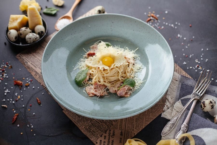 Food Fotograf Ein mit geriebenem Käse und einem Wachteleigelb belegtes Nudelgericht, serviert auf einem hellgrünen Teller. Der Teller steht auf einer Oberfläche, auf der verstreute Zutaten wie Wachteleier, Käsestücke und Gewürze verteilt sind. Daneben liegen eine Gabel und ein Messer. Essen fotografieren