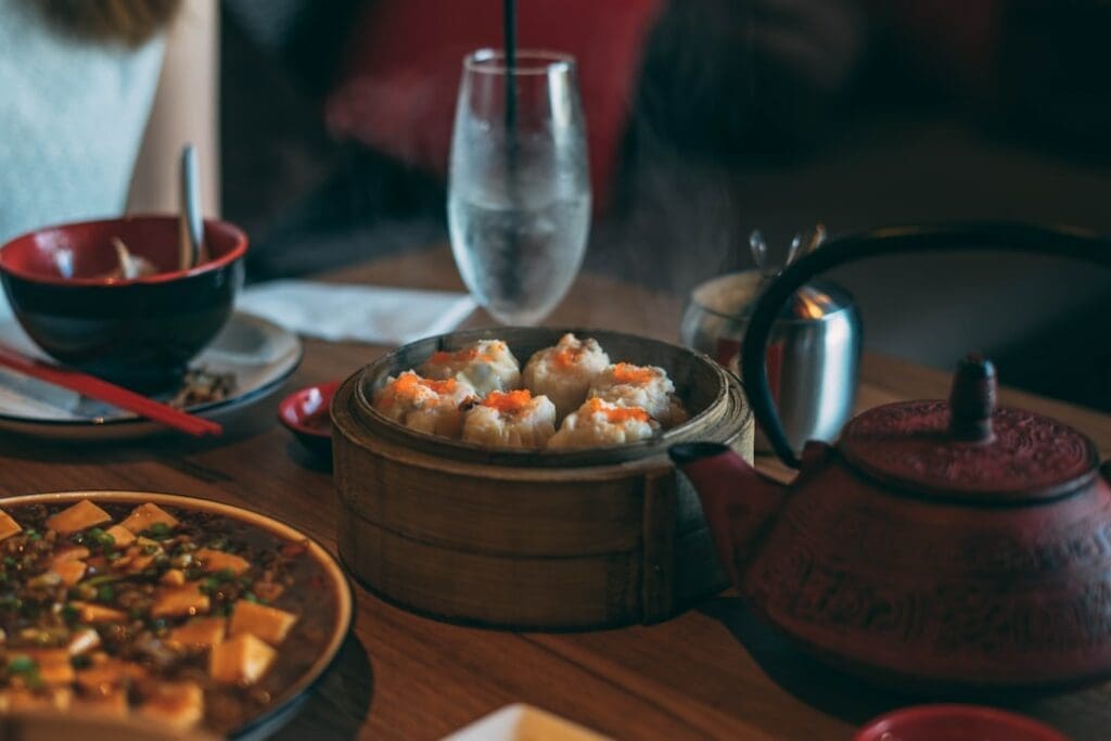 Der Food Fotograf - Ein gedeckter Tisch mit verschiedenen Gerichten, darunter ein Bambusdämpfer mit mit Rogen belegten Knödeln, eine rote Teekanne, ein Glas Wasser mit Strohhalm, eine Schüssel Suppe und ein Teller mit Essen. Von den Knödeln steigt Dampf auf, was darauf schließen lässt, dass sie frisch gekocht sind. Lecker Essen fotografieren.