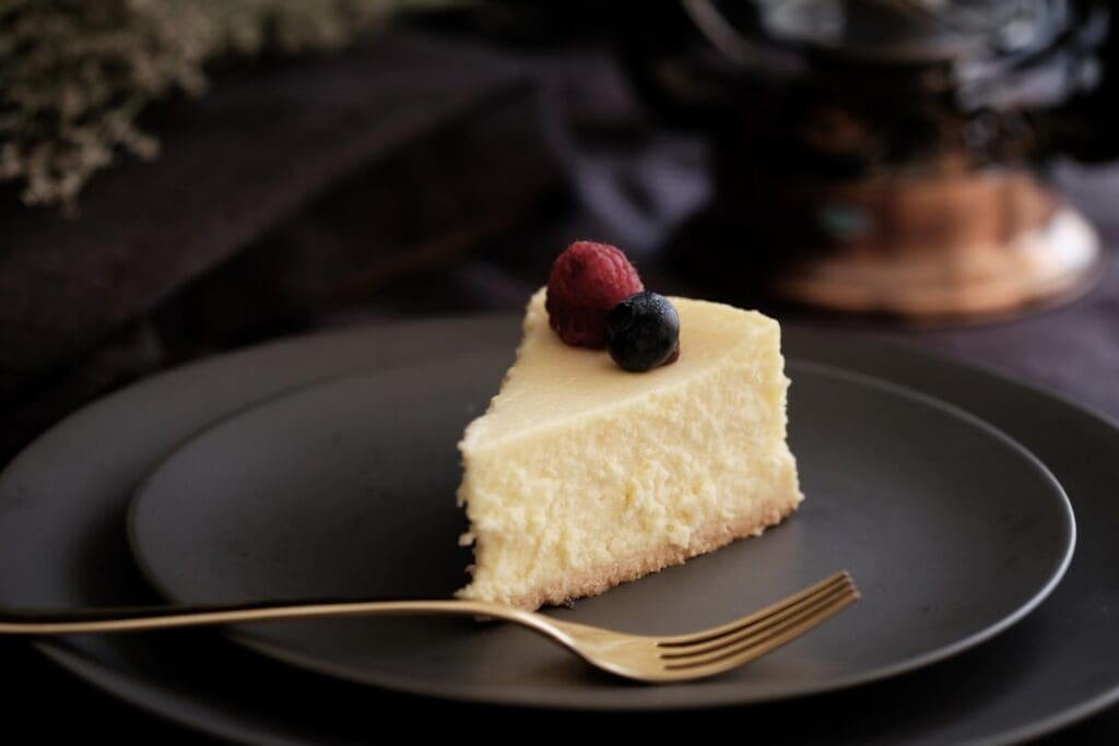 Food Fotograf Ein Stück Käsekuchen auf einem dunklen Teller, garniert mit einer einzelnen Himbeere und Blaubeere, dazu eine goldene Gabel. Der Hintergrund ist dunkel und leicht verschwommen, was den Fokus auf das Dessert verstärkt. Essen fotografieren