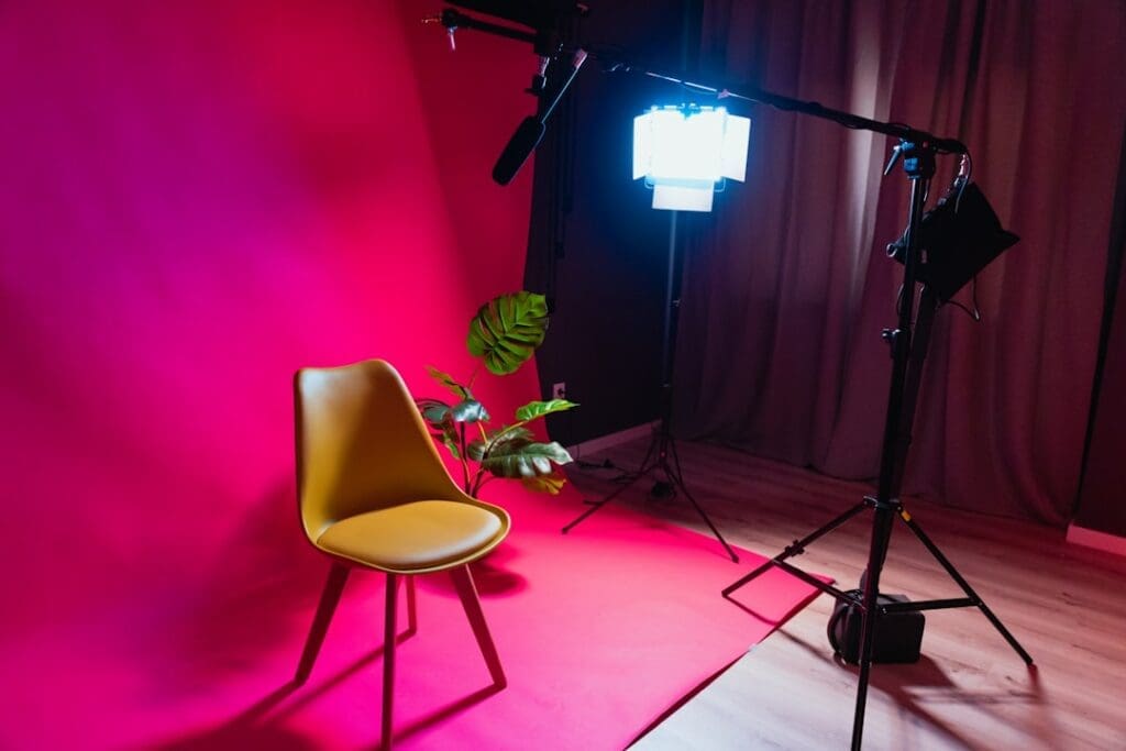 Der Food Fotograf - Eine Fotostudioszene, die einen gelben Stuhl auf einem roten Hintergrund mit Beleuchtungsausrüstung und einem Mikrofonaufbau zeigt. Links vom Stuhl befinden sich außerdem einige grüne Pflanzen. Der Boden ist aus Holz und im Hintergrund auf der rechten Seite befindet sich ein Vorhang. Lecker Essen fotografieren.