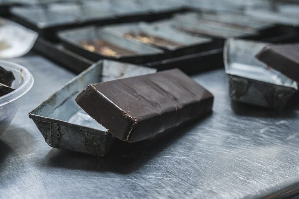 Der Food Fotograf - Eine Tafel dunkler Schokolade wird auf einer Edelstahloberfläche teilweise aus einer Metallform ausgepackt. Im Hintergrund sind weitere Schokoladenformen zu sehen. Lecker Essen fotografieren.