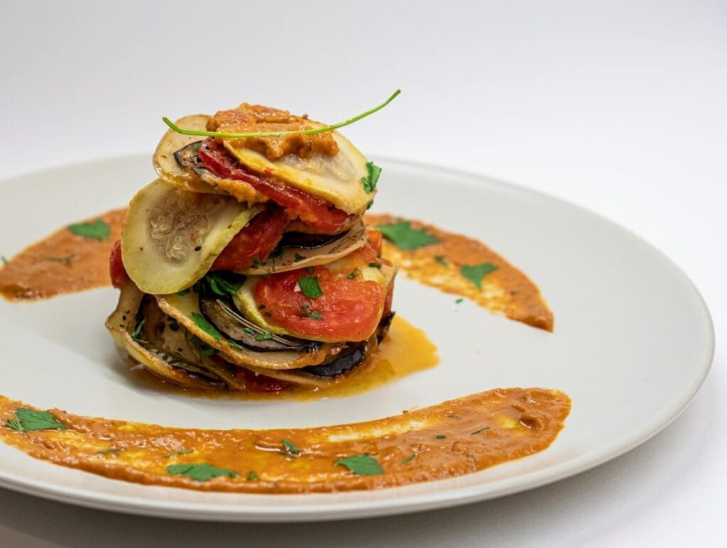 Der Food Fotograf - Eine Portion Ratatouille wird auf einem weißen Teller angerichtet. Die Ratatouille besteht aus ordentlich gestapelten Scheiben verschiedener Gemüsesorten, garniert mit einem grünen Zweig. Eine mit Kräutern garnierte Tomatensauce umgibt den Stapel auf dem Teller. Lecker Essen fotografieren.
