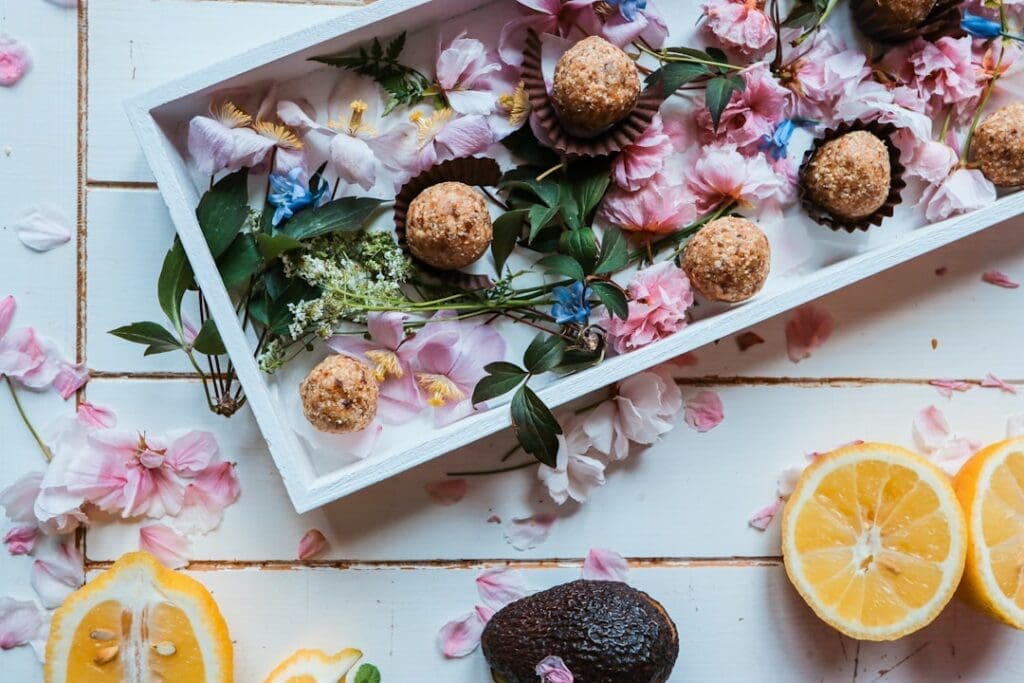 Food Fotograf Ein weißes, rechteckiges Tablett mit runden Süßigkeiten ist mit rosa Blütenblättern, grünen Blättern und anderen floralen Elementen geschmückt. Um das Tablett herum auf dem Tisch liegen halbierte Zitronen, eine Avocado und weitere verstreute rosa Blütenblätter und Blätter. Essen fotografieren