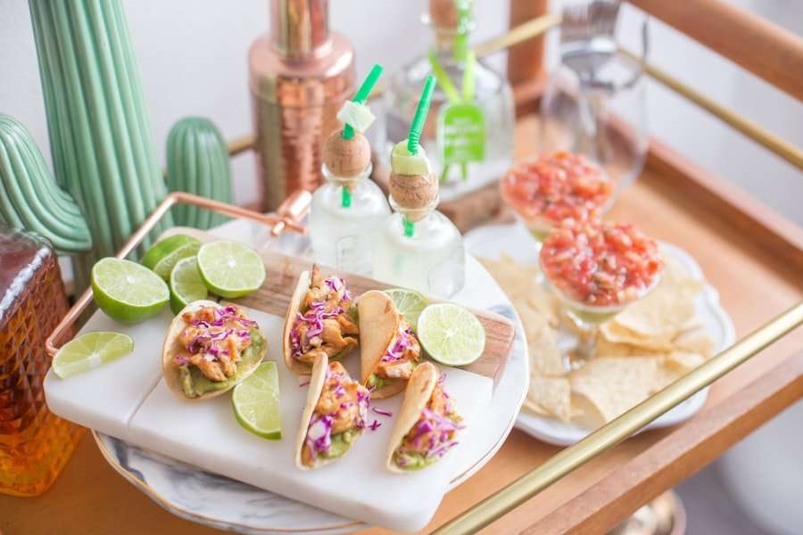 Der Food Fotograf - Auf einem kleinen Servierwagen aus Holz steht ein Tablett mit Mini-Tacos, garniert mit Limettenspalten und geraspeltem Gemüse. Daneben stehen zwei Gläser mit Salsa und Tortillachips. Im Hintergrund sind Flaschen und Gläser mit grünen Strohhalmen ausgestellt. Lecker Essen fotografieren.