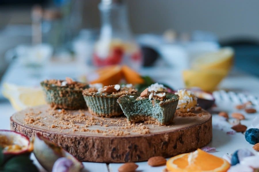 Food Fotograf Eine Nahaufnahme eines Holzbretts, auf dem drei grünlich-braune Cupcakes mit gehackten Mandeln liegen. Aus einem Cupcake ist ein Biss herausgeschnitten. Im Hintergrund sind verschwommene Elemente von Früchten und anderen Esswaren zu sehen, die auf einer weißen Oberfläche verstreut sind. Essen fotografieren