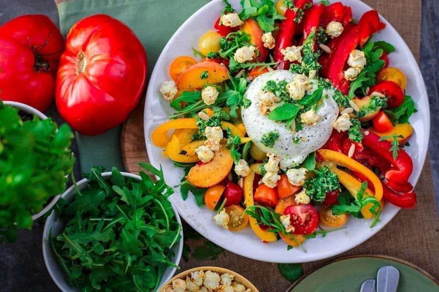 Food Fotograf Ein bunter Salat auf einem weißen Teller besteht aus Burrata-Käse, Rucola, roten und gelben Paprikaschoten, Kirschtomaten und Popcorn. Neben dem Teller stehen zwei Tomaten und eine Schüssel Rucola. Eine grüne Serviette und Besteck sind teilweise sichtbar. Essen fotografieren