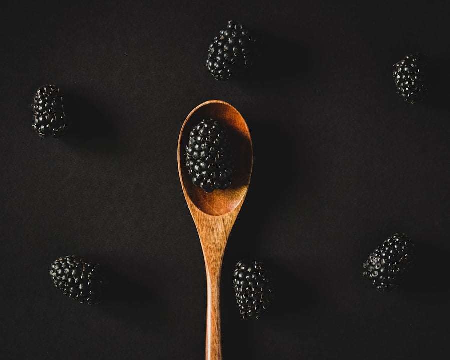 Der Food Fotograf - Auf einem Holzlöffel liegt eine einzelne Brombeere, umgeben von fünf weiteren Brombeeren, die auf einem schwarzen Hintergrund verstreut sind. Die Brombeeren sind ungleichmäßig verteilt, wodurch eine einfache und minimalistische visuelle Komposition entsteht. Lecker Essen fotografieren.