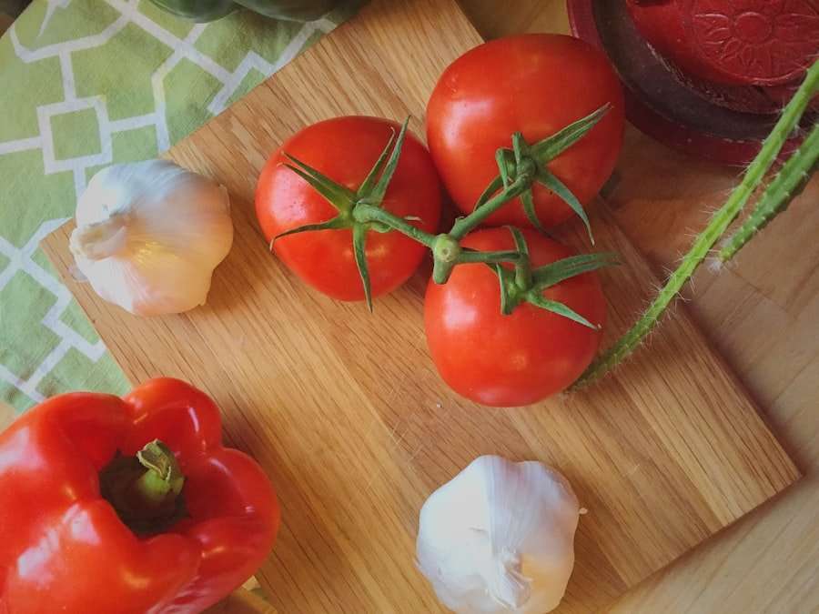 Food Fotograf Hannover - Drei rote Tomaten an einer Ranke, zwei Knoblauchknollen, eine rote Paprika und eine grüne Artischocke sind auf einem hölzernen Schneidebrett angeordnet. Im Hintergrund ist ein Teil eines grün gemusterten Tuchs zu sehen. Food Fotografie Hannover.