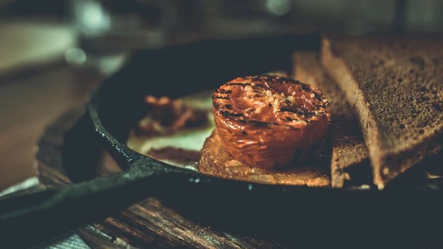 Der Food Fotograf - Eine gegrillte halbe Tomate liegt zusammen mit zwei weiteren Brotscheiben auf einer Brotscheibe in einer Pfanne. Das Essen ist auf einer Holzoberfläche angerichtet und der Hintergrund ist unscharf. Lecker Essen fotografieren.