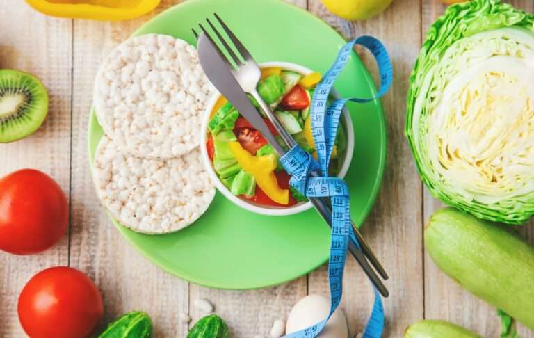 Food Fotograf Hannover - Ein grüner Teller mit zwei Reiskuchen, einer Schüssel Salat und Besteck, das mit einem blauen Maßband umwickelt ist. Um den Teller herum liegen frische Gemüsesorten wie Tomaten, Gurken, Zucchini, Kohl, Kiwi und eine gelbe Paprika. Wunderschön aufgenommen von Food Fotograf Hannover. Lecker Essen fotografieren.