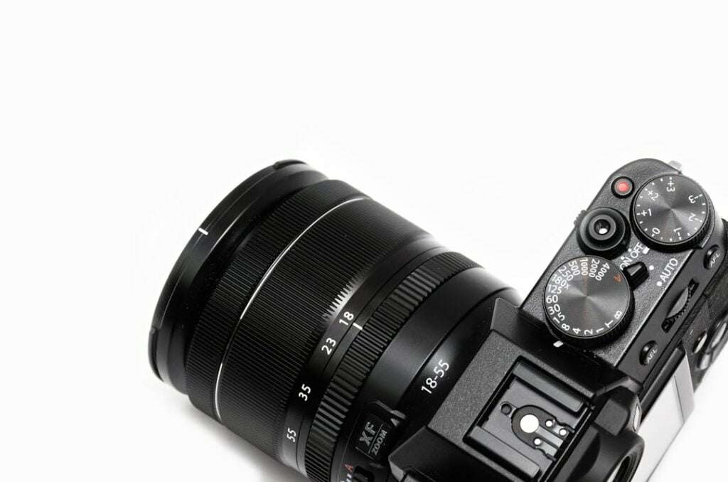 Der Food Fotograf - Eine Nahaufnahme einer schwarzen Digitalkamera, auf der das Objektiv und die Einstellräder zu sehen sind. Das Objektiv ist mit Brennweiten von 18 bis 55 mm gekennzeichnet. Die Einstellräder oben enthalten Einstellungen für verschiedene Fotomodi und Anpassungen. Der Hintergrund ist weiß. Lecker Essen fotografieren.