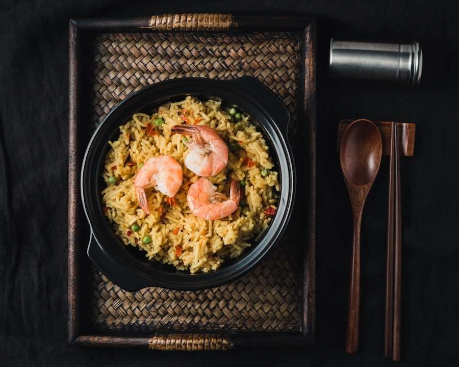 Der Food Fotograf - Eine schwarze Schüssel mit gebratenem Garnelenreis steht auf einem geflochtenen Tablett. Der Reis ist mit drei Garnelen garniert. Rechts vom Tablett liegen ein Holzlöffel, ein Paar Essstäbchen und ein kleiner zylindrischer Behälter. Der Hintergrund ist dunkel. Lecker Essen fotografieren.