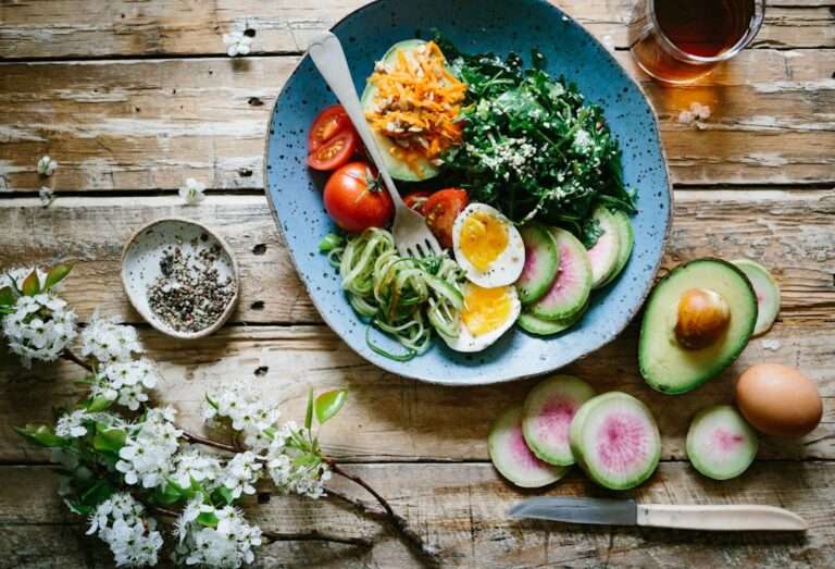 Food Fotograf Hannover - Auf einem rustikalen Holztisch steht ein blauer Teller mit einer Mischung aus Salatgemüse, darunter Blattgemüse, Zucchini-Nudeln, Avocadoscheiben, halbierte Kirschtomaten, hartgekochte Eier und Radieschenscheiben. Ein Glas Tee und ein Zweig mit weißen Blüten sind ebenfalls vorhanden. Lecker Essen fotografieren.