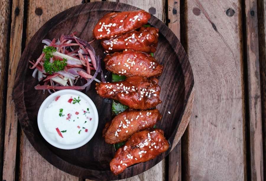 Food Fotograf Hannover - Ein Holzteller mit sechs glasierten Hähnchenflügeln, garniert mit Sesamkörnern, eingelegten Zwiebeln und einem Schälchen weißer Dip-Sauce, garniert mit Kräutern und roten Stückchen. Darunter ist die hölzerne Tischoberfläche zu sehen. Food Fotografie Hannover.