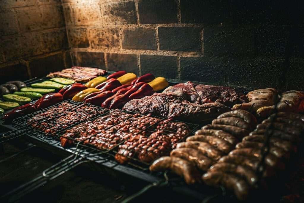Food Fotograf Hannover - Verschiedene Fleischsorten, Wurst und Gemüse, darunter Paprika und Zucchini, werden über offener Flamme gegrillt. Das Essen ist ordentlich auf Grillrosten vor einem Backsteinmauerhintergrund angeordnet. Die gedämpfte Beleuchtung unterstreicht die Kunstfertigkeit der Lebensmittelfotografie. Food Fotografie Hannover.
