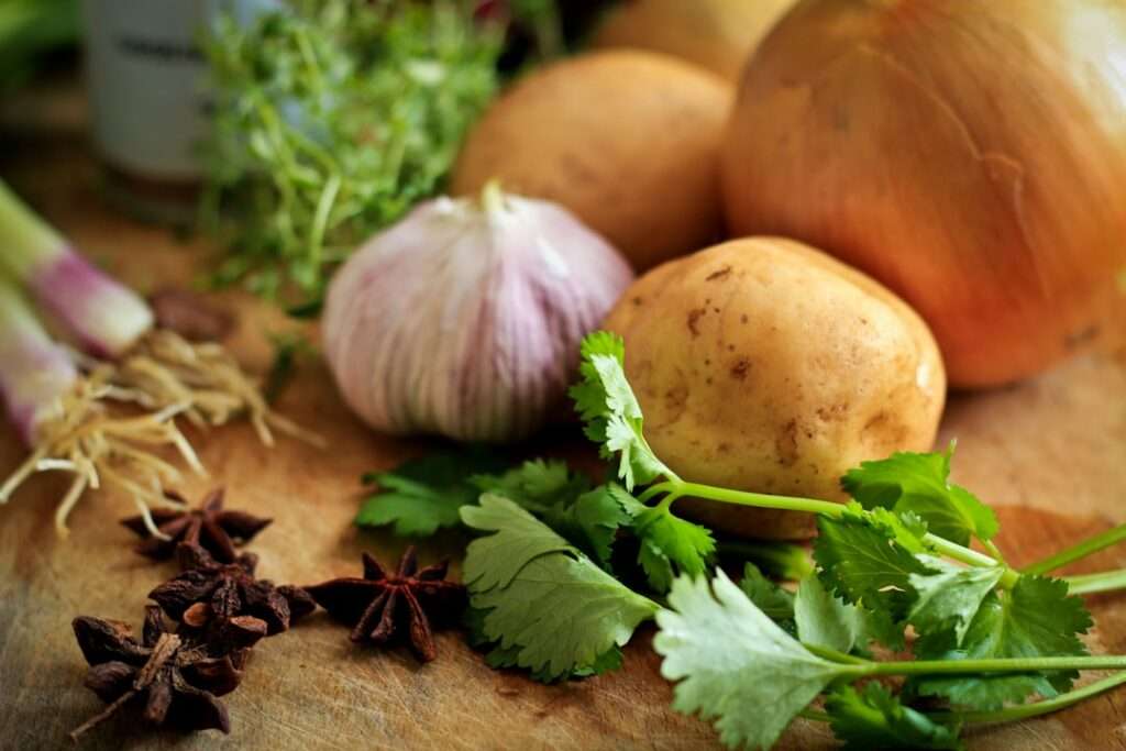 Food Fotograf Hannover - Eine Nahaufnahme verschiedener Gemüsesorten und Kräuter auf einer Holzoberfläche, darunter eine Kartoffel, eine Zwiebel, eine Knoblauchknolle, Sternanis und Koriander. Food Fotografie Hannover.