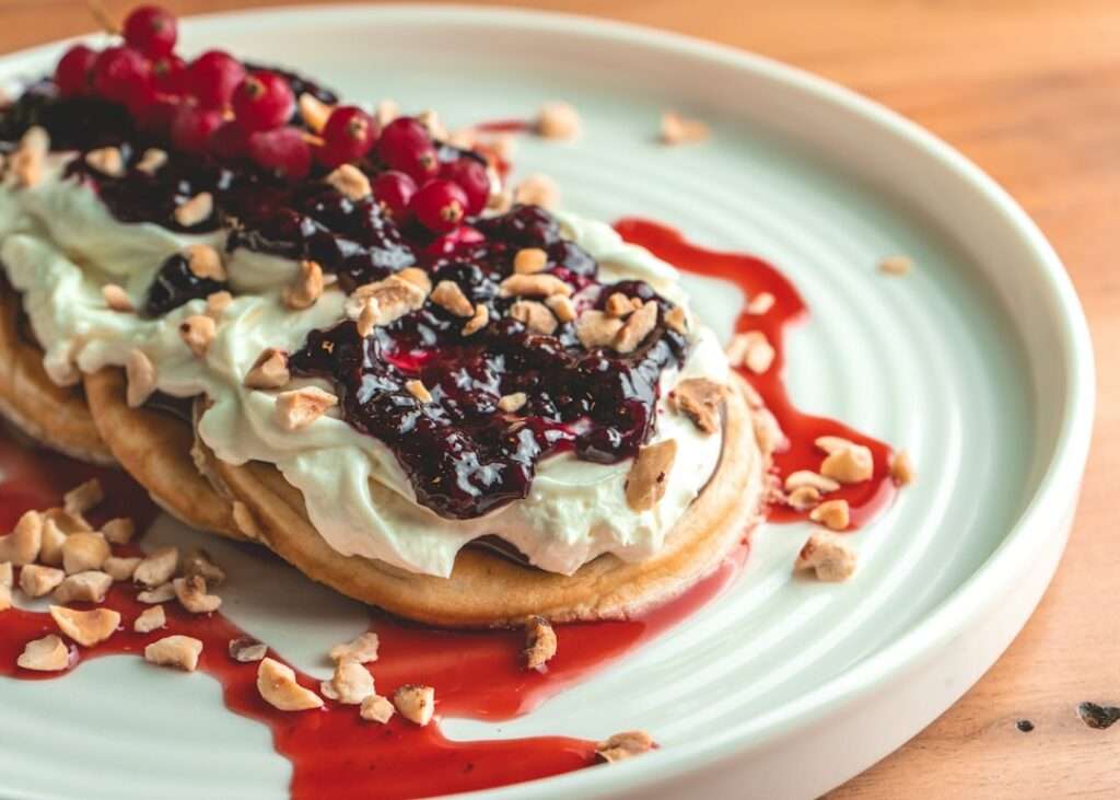Der Food Fotograf - Auf einem Teller liegt ein Dessert mit Gebäck, das mit Schlagsahne und dunkler Beerensauce belegt und mit gehackten Nüssen bestreut ist. Obenauf liegen rote Beeren an einem Stiel. Das Gericht steht auf einem weißen Teller auf einer Holzoberfläche. Lecker Essen fotografieren.