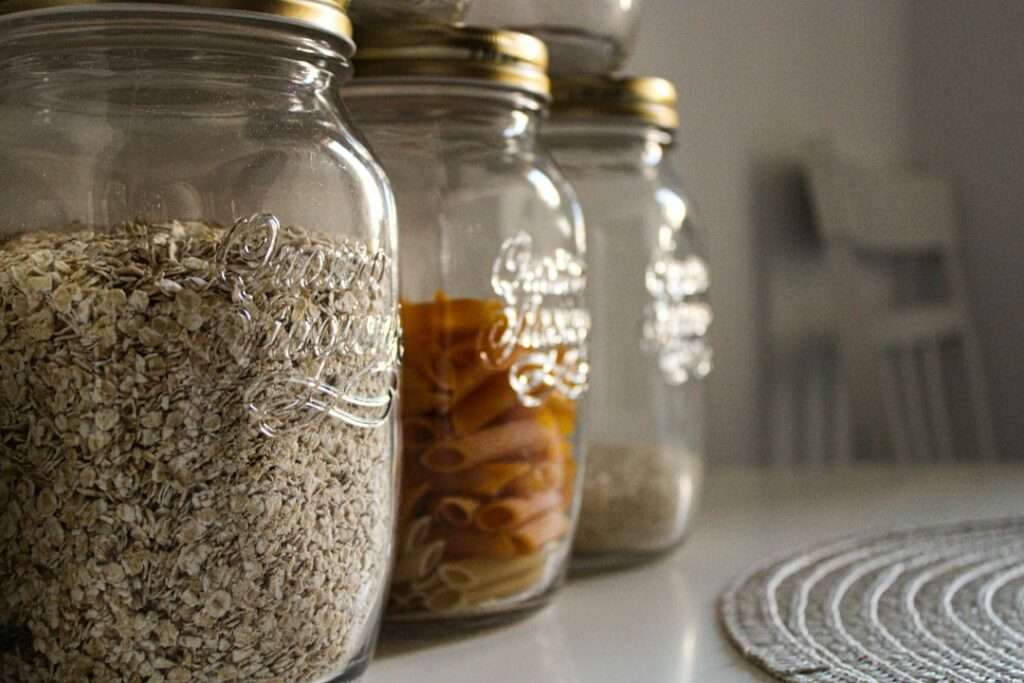 Food Fotograf Hannover - Vier Gläser mit Metalldeckeln stehen in einer Reihe auf einer weißen Fläche und präsentieren Hafer, Nudeln und andere Getreidesorten. Auf der rechten Seite ist teilweise eine runde gewebte Matte zu sehen. Die Food-Fotografie fängt eine harmonische Szene mit einem verschwommenen Stuhl und Tisch im Hintergrund ein. Food Fotografie Hannover.