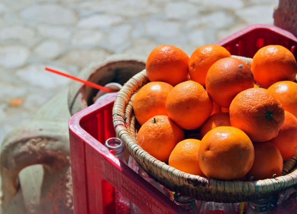 Food Fotograf Hannover - Ein mit Orangen gefüllter geflochtener Korb steht auf einer umgedrehten roten Plastikkiste. Im Hintergrund ist ein verschwommener Steinboden zu sehen, aus dessen linker Seite ein roter Strohhalm herausragt und auf der linken Seite teilweise eine Steinvase zu sehen ist. Food Fotografie Hannover.