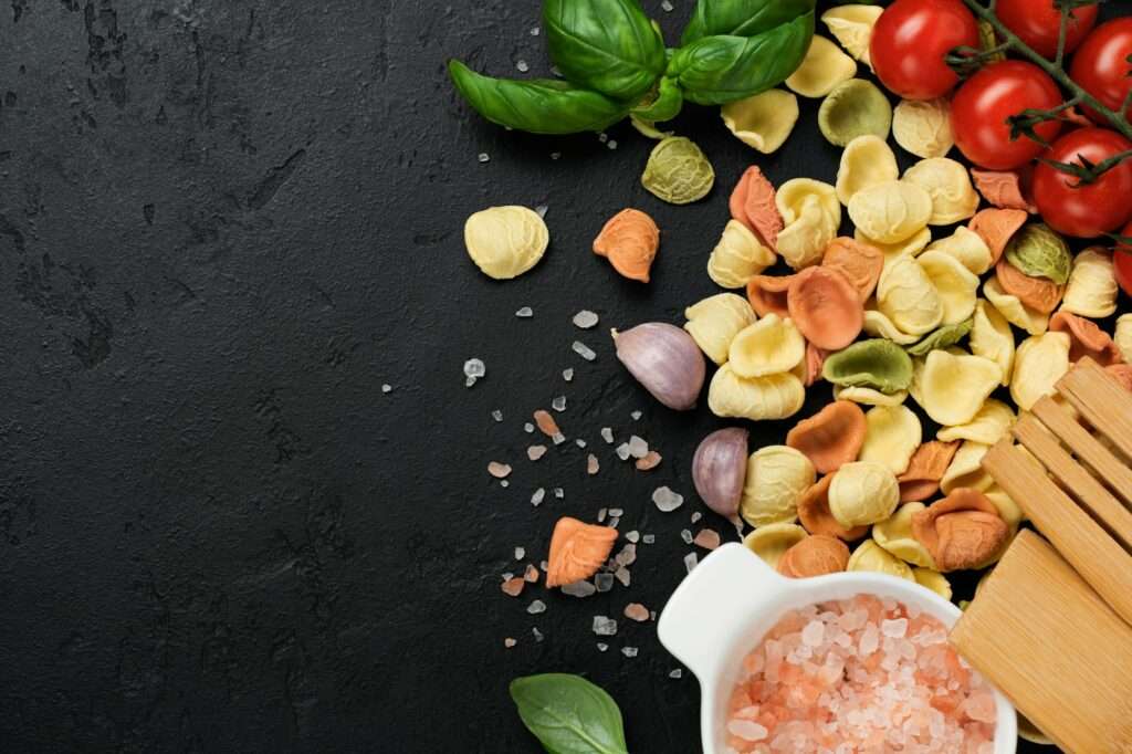 Food Fotograf Hannover - Eine Auswahl trockener, bunter Tortellini, Kirschtomaten, Knoblauchzehen, frische Basilikumblätter und ein Holzlöffel sind auf einer dunklen strukturierten Oberfläche ausgebreitet. Außerdem ist eine weiße Schüssel mit rosa Himalaya-Salz zu sehen. Food Fotografie Hannover.