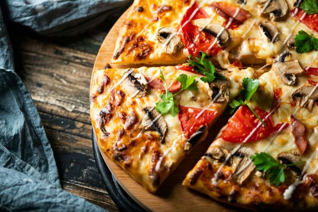 Food Fotograf Hannover - Eine Nahaufnahme einer geschnittenen Pizza auf einem hölzernen Servierbrett. Die Pizza ist mit geschmolzenem Käse, Pilzen, Tomatenscheiben und Speck belegt und mit frischer Petersilie garniert. Das Brett liegt auf einem rustikalen Holztisch, seitlich ist teilweise eine graue Serviette zu sehen. Food Fotografie Hannover.