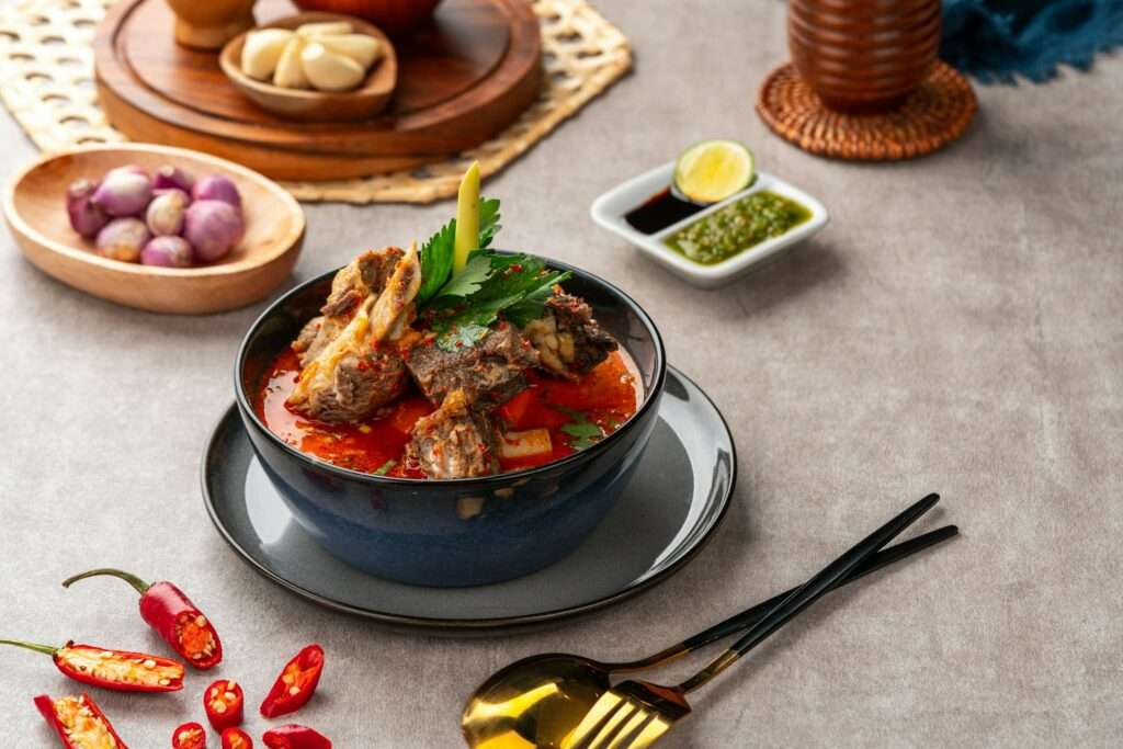 Food Fotograf Hannover - Eine Schüssel würzigen Rindfleischeintopfs, garniert mit Kräutern, steht auf einem Tisch und schafft eine einladende Szene für Food-Fotografie. Um die Schüssel herum befinden sich rote Chilischoten, geschälter Knoblauch, Schalotten, eine kleine Schale mit Soßen sowie eine Gabel und ein Löffel aus Metall. Die neutral gefärbte Oberfläche im Hintergrund verstärkt die leuchtenden Farben der Schale. Food Fotografie Hannover.