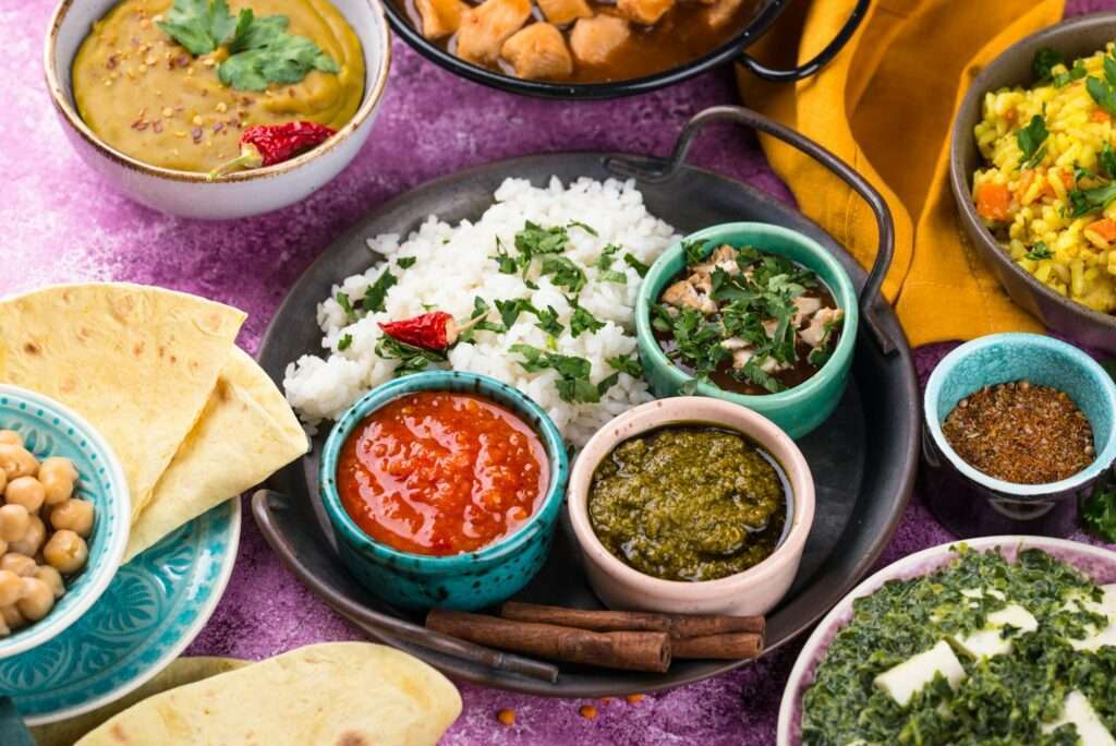 Food Fotograf Hannover - Eine Auswahl indischer Gerichte ist kunstvoll auf einem Tisch arrangiert, darunter Schüsseln mit Curry, Reis, Chutneys und Linsen mit Naan-Brot als Beilage. Die Speisenpräsentation auf bunten Tellern und Schüsseln vor einem violetten Untergrund ist perfekt für die „Nachbearbeitung von Essensfotos“. Food Fotografie Hannover.