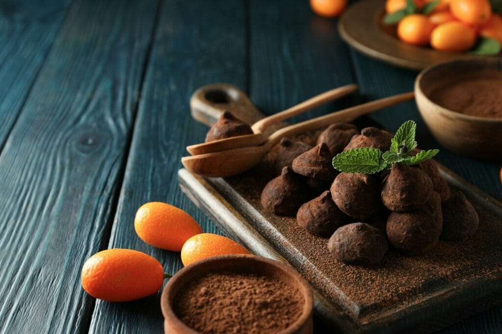 Food Fotograf Hannover - Auf einem Holzbrett werden mit Kakaopulver bestäubte Schokoladentrüffel präsentiert, elegant garniert mit Minzblättern. Daneben stehen frische Kumquats neben einer Schale Kakaopulver. Die Szene wird durch die richtige Beleuchtung in der Food-Fotografie vor einer dunkelblauen Holzoberfläche noch verstärkt. Food Fotografie Hannover.