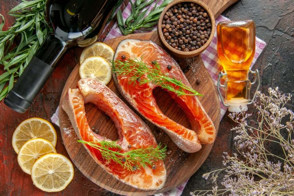 Food Fotograf Hannover - Zwei rohe Lachssteaks mit Dill liegen auf einem runden Holzschneidebrett, umgeben von Zitronenscheiben, einer Flasche Wein und einer kleinen Schale mit Pfefferkörnern. Eine kleine Flasche Öl und verstreute getrocknete Kräuter verleihen dem Food-Fotografie-Hintergrund Tiefe. Food Fotografie Hannover.