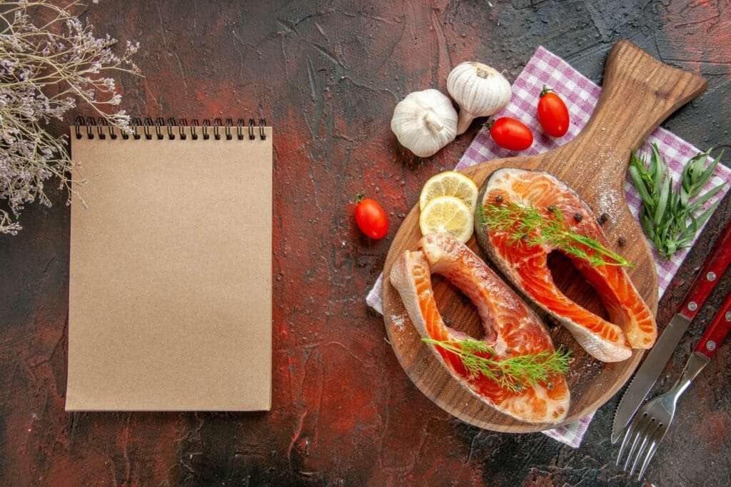 Food Fotograf Hannover - Auf einem Holzbrett liegen zwei rohe Lachssteaks, garniert mit Zitronenscheiben und Dill, und fangen einen perfekten Moment für eine Food-Fotografie ein. Tomaten, Knoblauch und Kräuter umgeben das Brett. Messer und Gabel liegen auf einem karierten Tuch, während ein offenes leeres Notizbuch auf einer dunklen Oberfläche an diesem gemütlichen Ort auf Inspiration wartet. Food Fotografie Hannover.